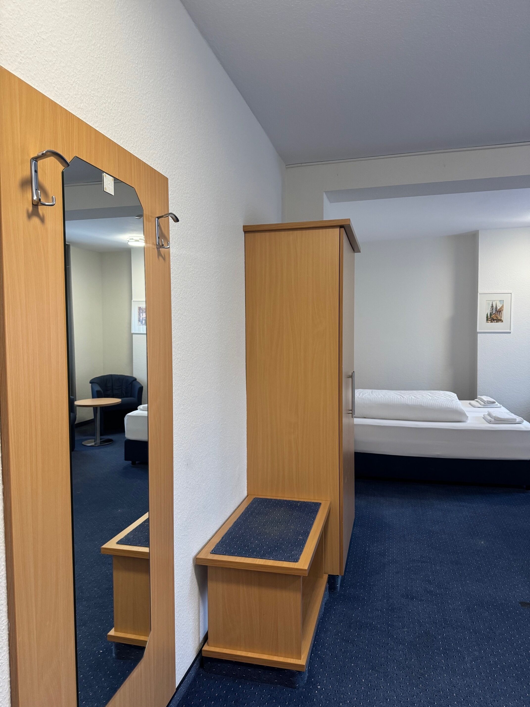 A hotel room with a wooden wardrobe, a small wooden table, a bed, and a mirror.