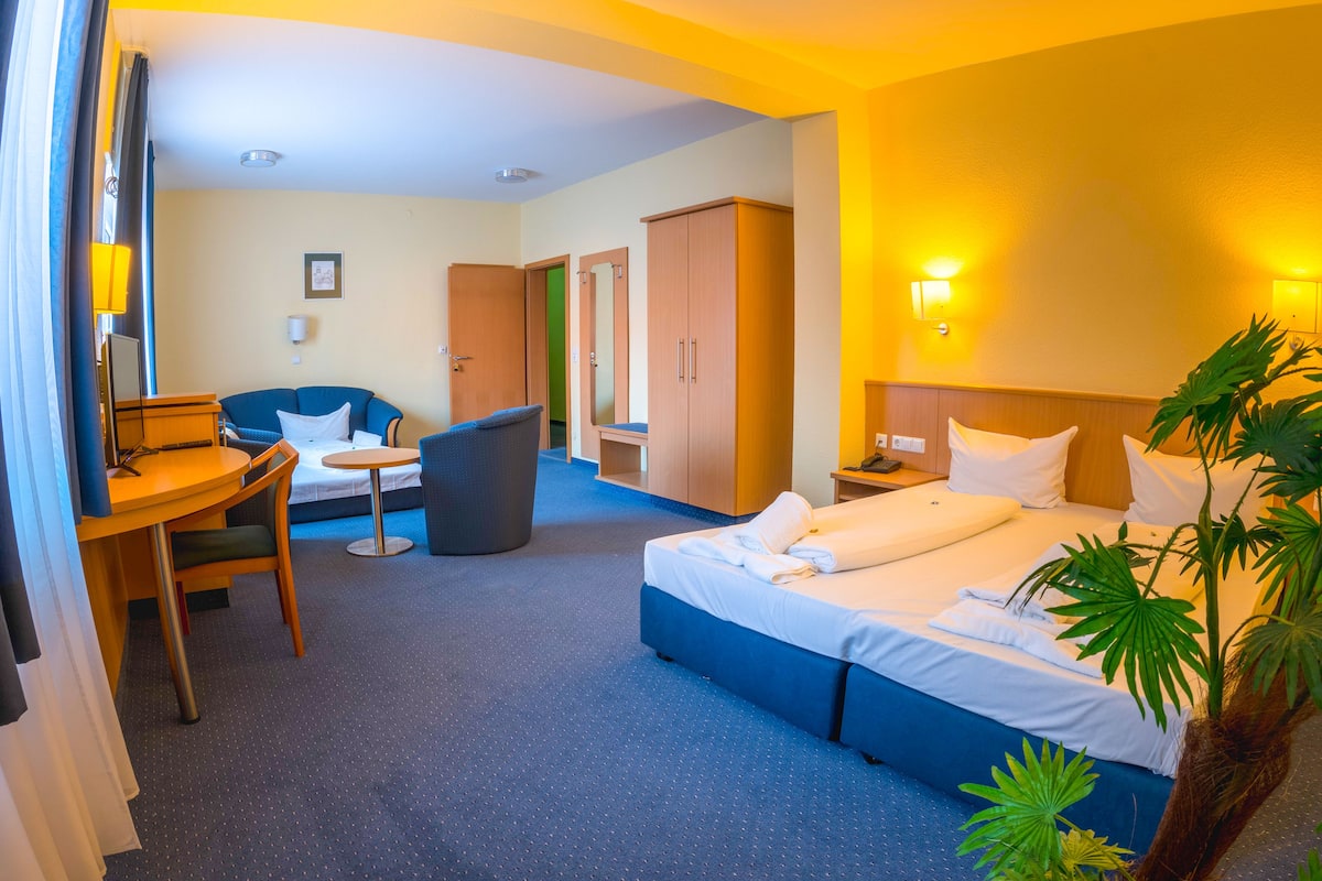 Una habitación de hotel con cama, escritorio, silla, mesita, cubo de basura y armario.
