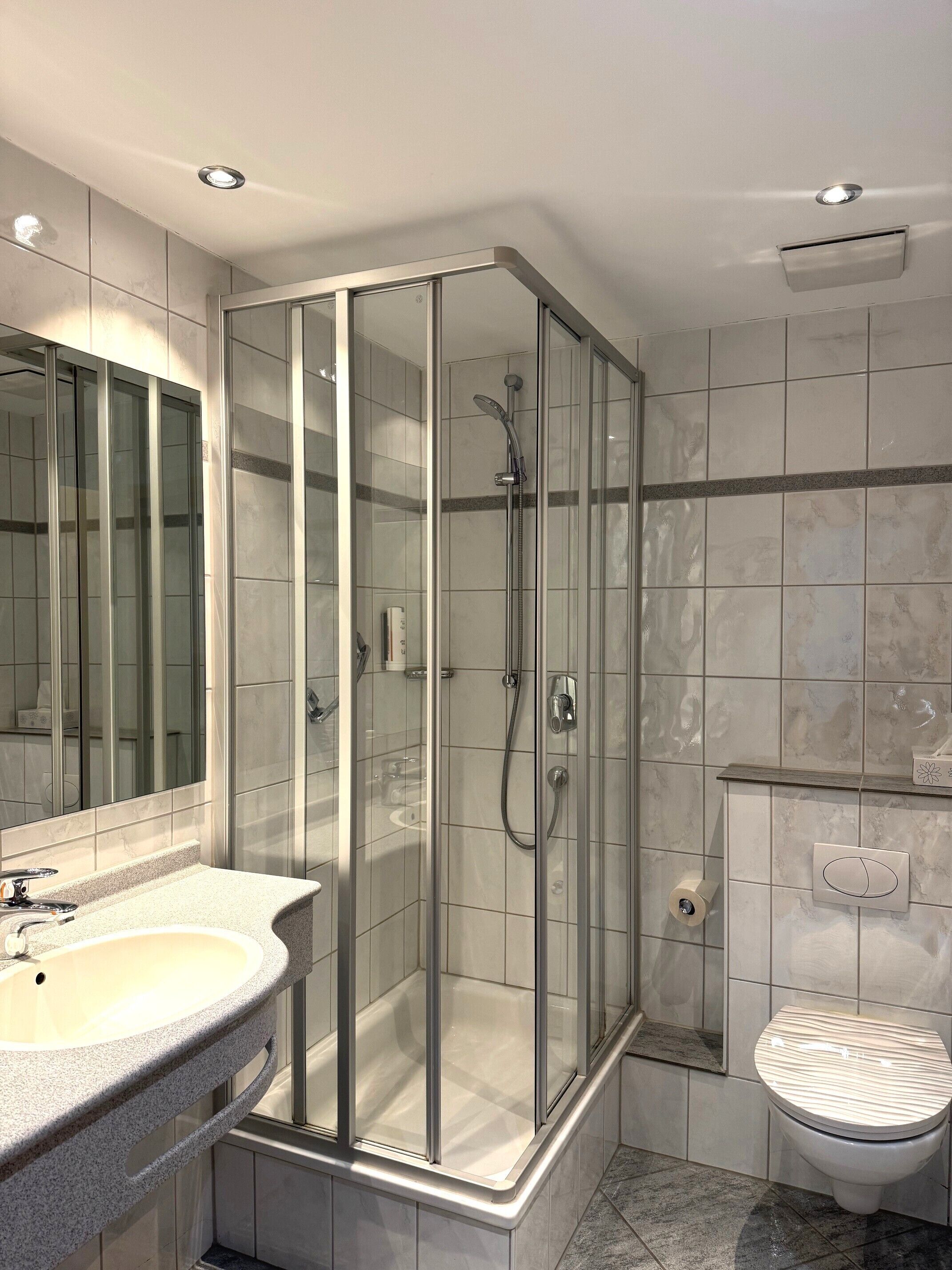 A modern bathroom with a glass-enclosed shower, a sink with a marble countertop, and a toilet with a marble lid.