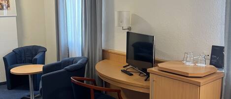 Habitación de hotel con escritorio, dos sillas, una mesita, televisor y minibar.