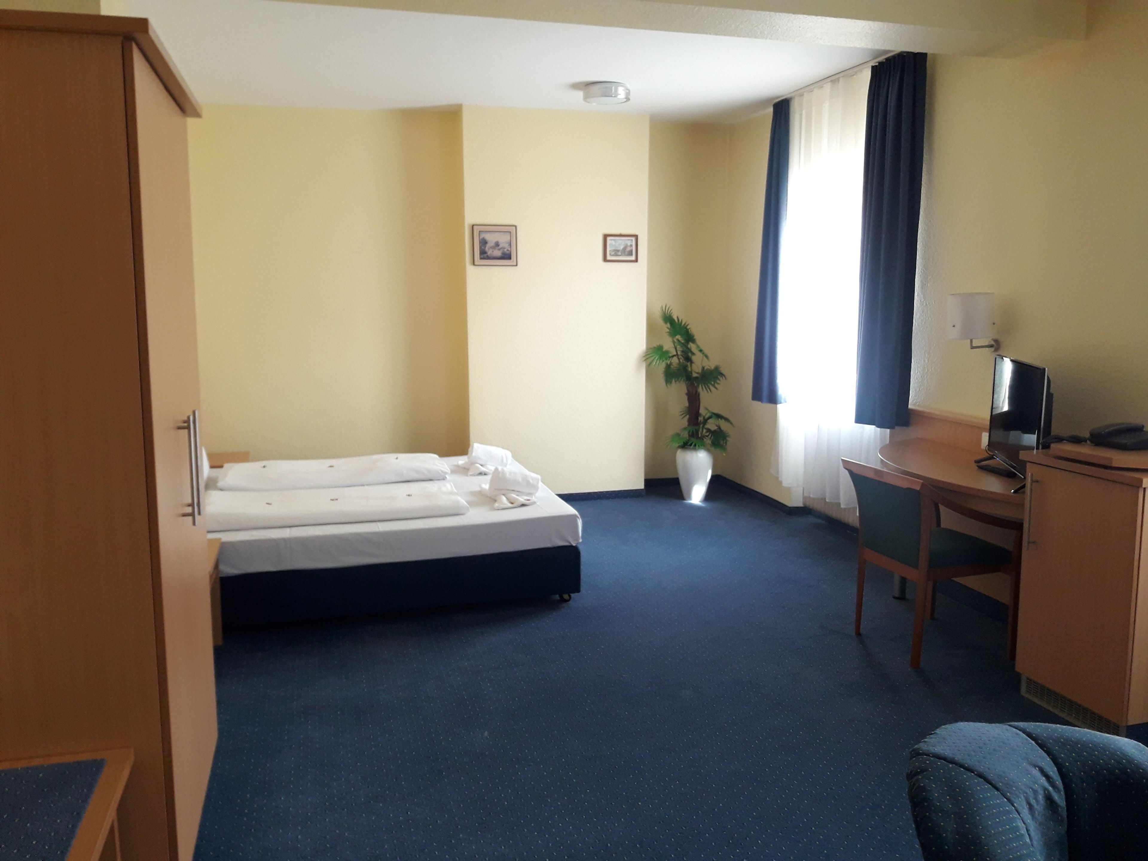 Ein Hotelzimmer mit einem Bett, einem Schreibtisch mit Computer, einem Stuhl, einer Zimmerpflanze und zwei gerahmten Bildern an der Wand.