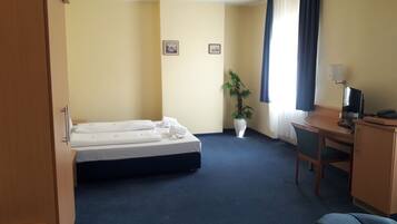 Ein Hotelzimmer mit einem Bett, einem Schreibtisch mit Computer, einem Stuhl, einer Zimmerpflanze und zwei gerahmten Bildern an der Wand.