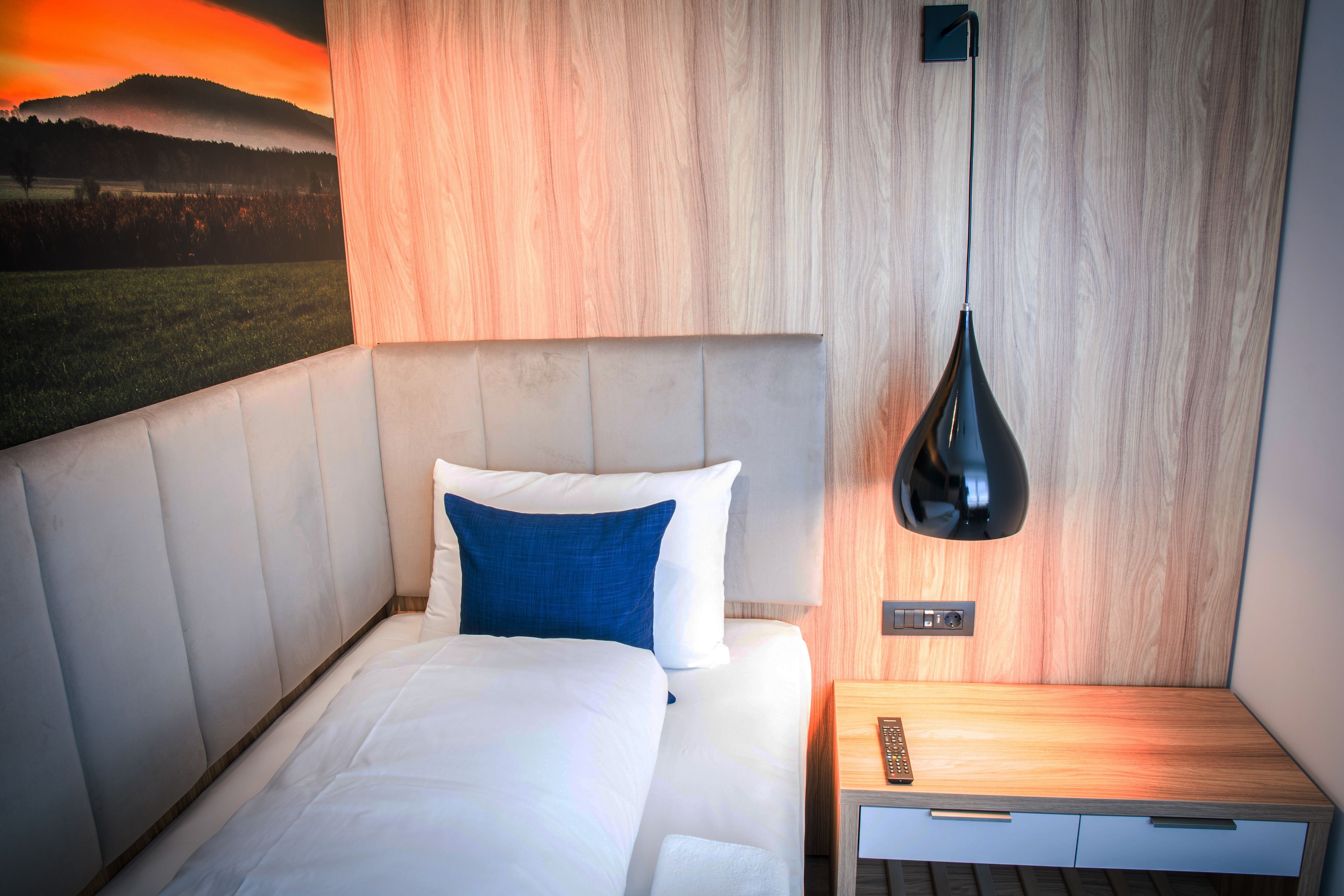 A modern hotel room with a bed, a nightstand, a wall-mounted lamp, and a painting of a landscape.