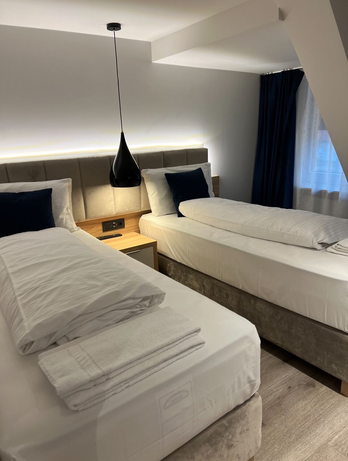 A modern hotel room with two beds, a nightstand, a lamp, and a window with curtains.