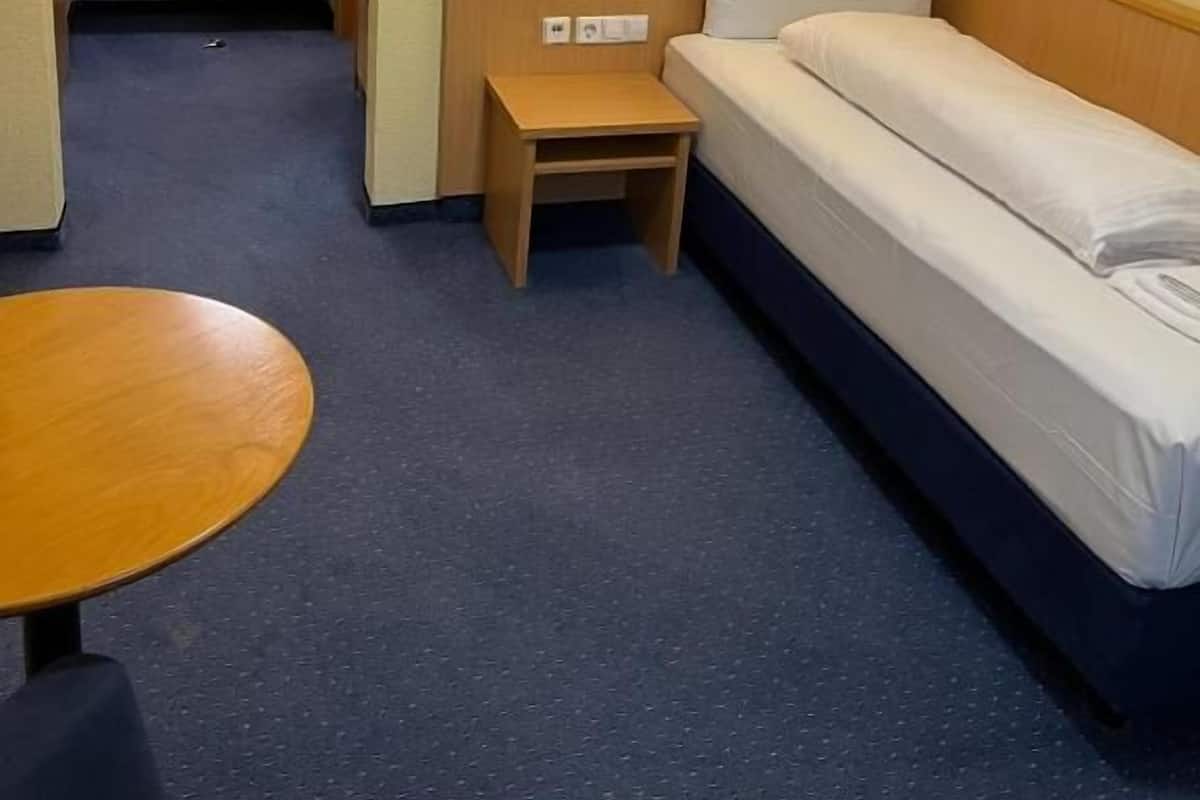 Habitación de hotel pequeña con una cama individual, una mesita de noche de madera, una lámpara fijada a la pared y una mesa redonda.