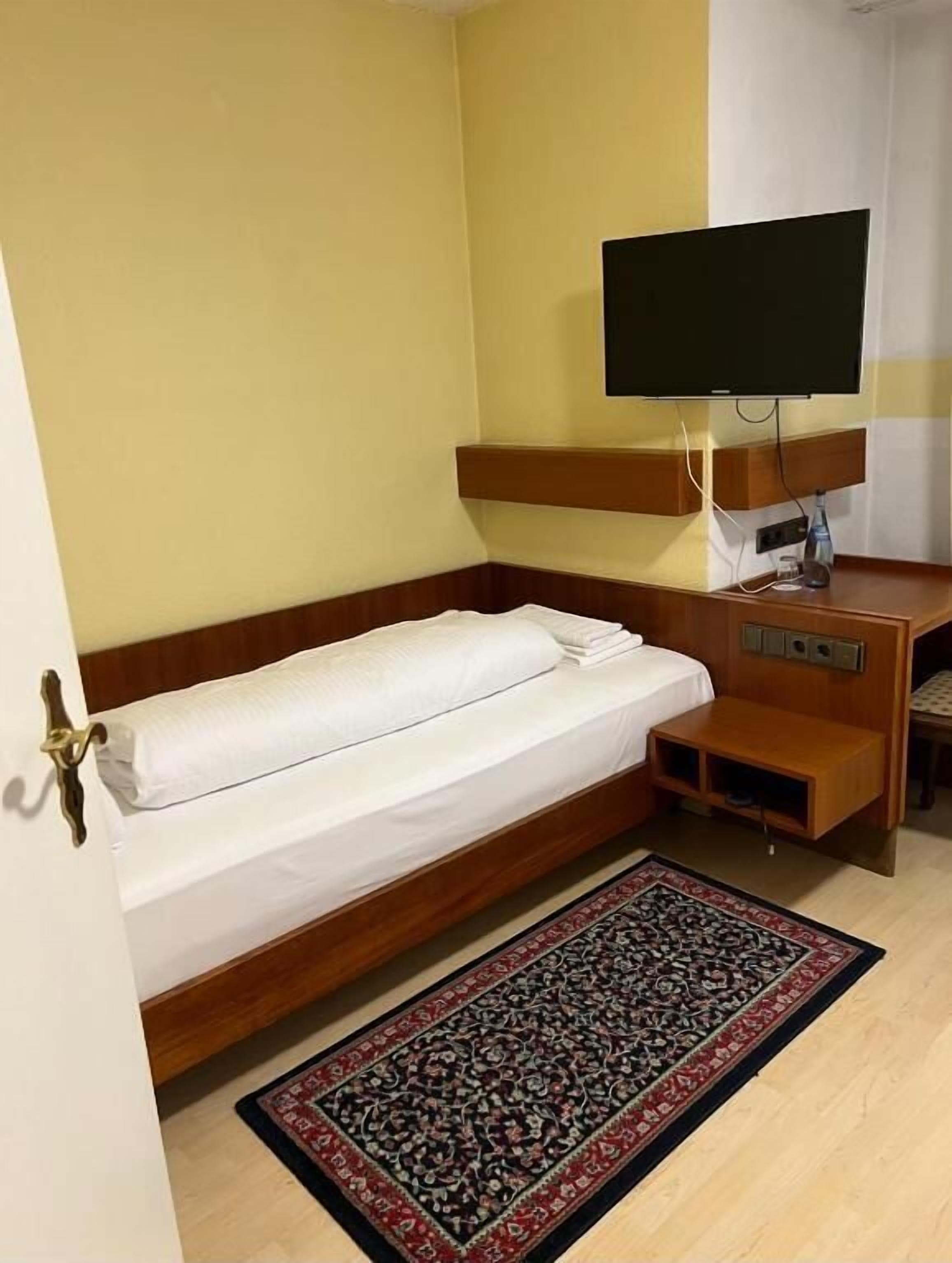 A single bed with a wooden headboard, a bedside table, a television mounted on the wall, and a patterned rug on the floor.
