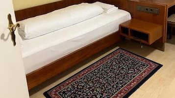 A single bed with a wooden headboard, a bedside table, a television mounted on the wall, and a patterned rug on the floor.