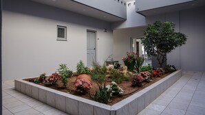Un edificio moderno con techo plano, un pequeño jardín con plantas y flores diversas, y piso de baldosas.