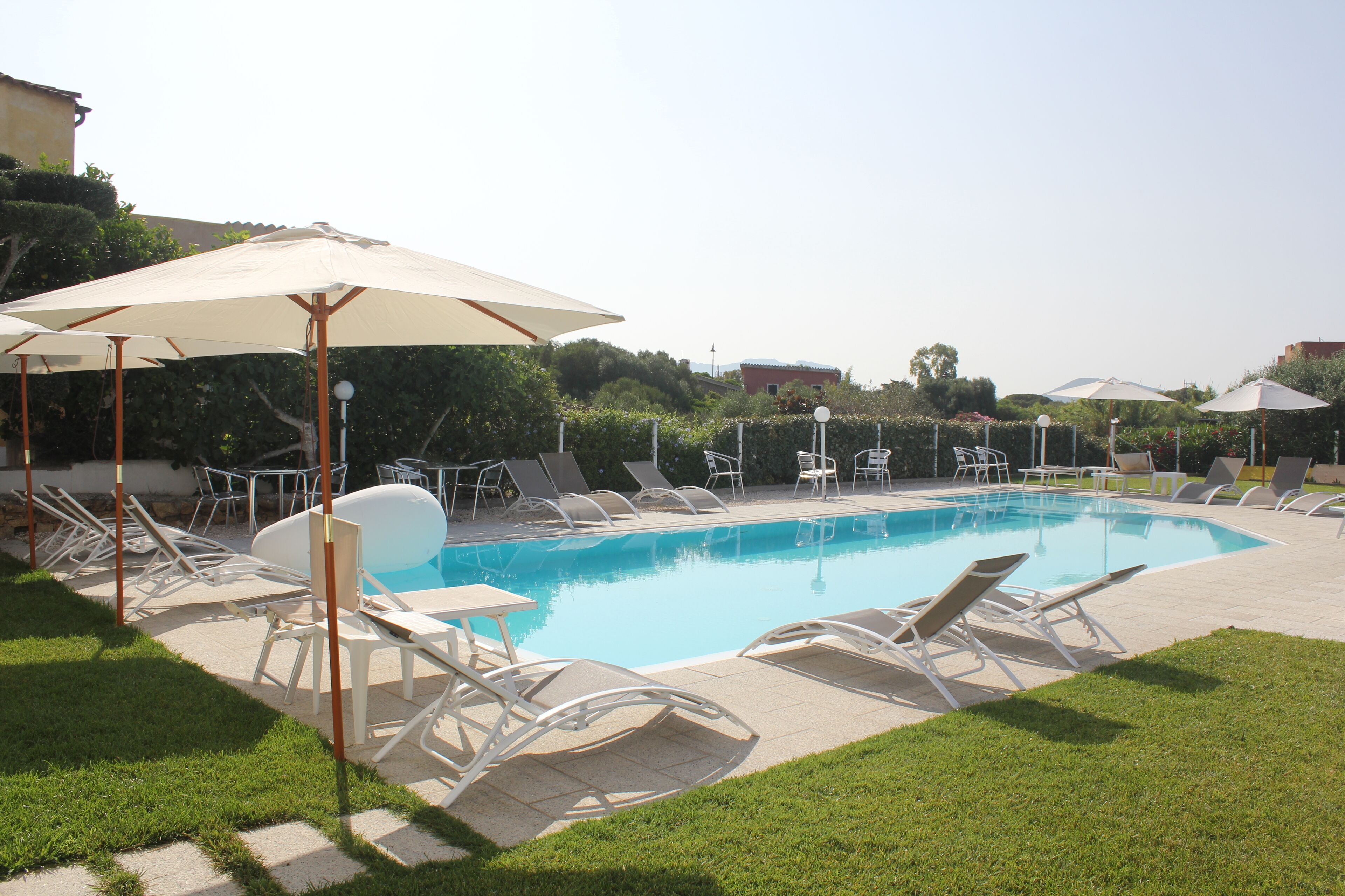 A swimming pool with lounge chairs and umbrellas, surrounded by greenery and buildings in the background.