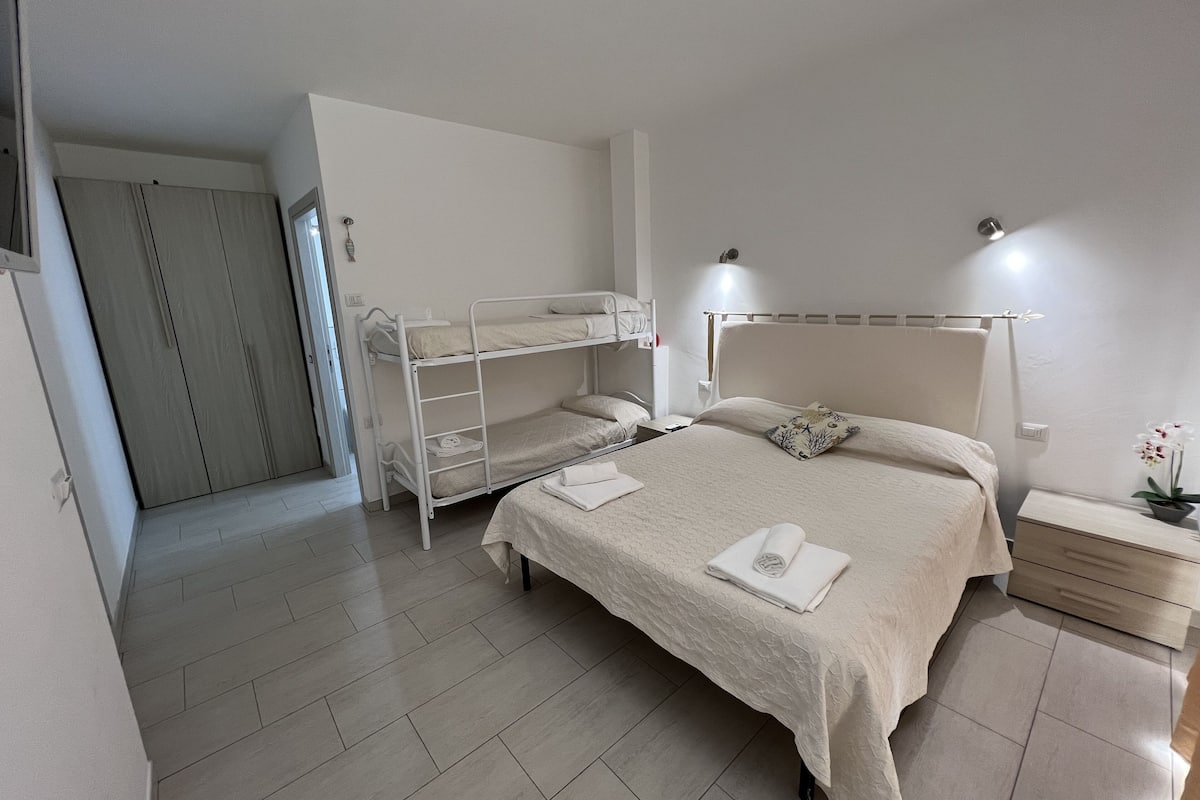 A bedroom with a bunk bed, a single bed, a bedside table with a lamp, and a wardrobe.