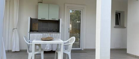 A covered outdoor dining area with a table and chairs, a kitchen in the background, and a door leading to a glass door.