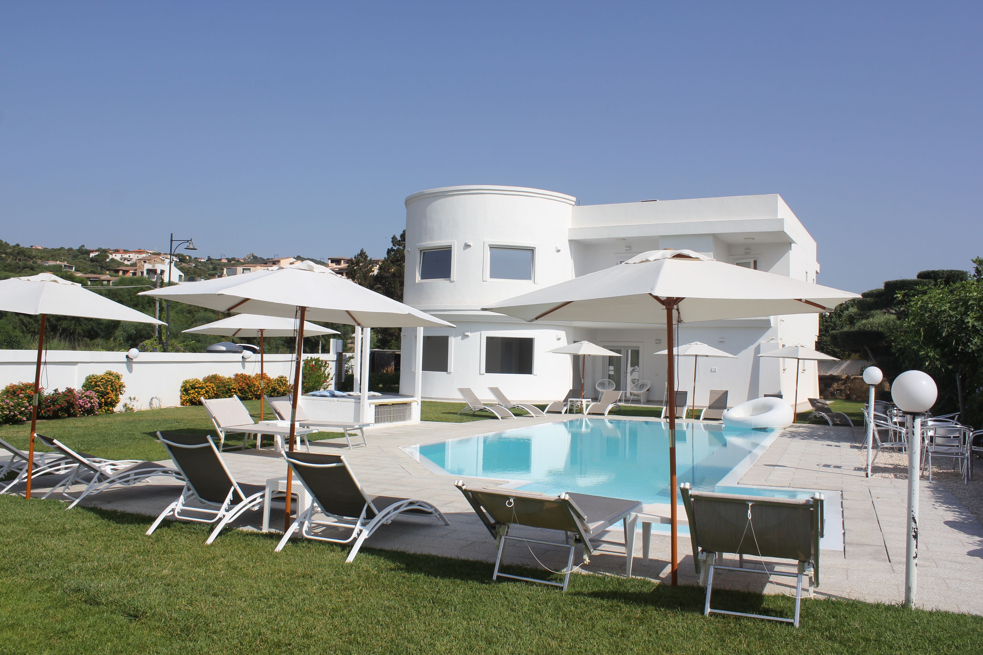 Un espace piscine moderne avec des chaises longues et des parasols, une piscine à l’eau d’un bleu limpide et un bâtiment blanc en arrière-plan.