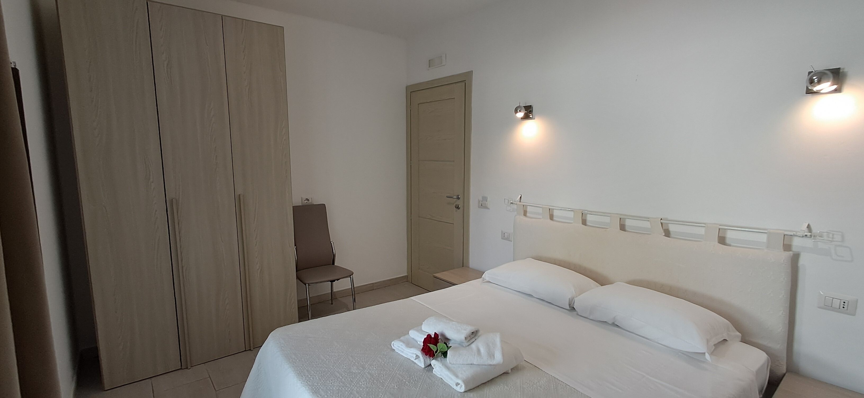 Hotellihuone, jossa on suuri vaatekaappi, yksi sänky valkoisilla vuodevaatteilla, tuoli ja kaksi seinään kiinnitettyä valaisinta.