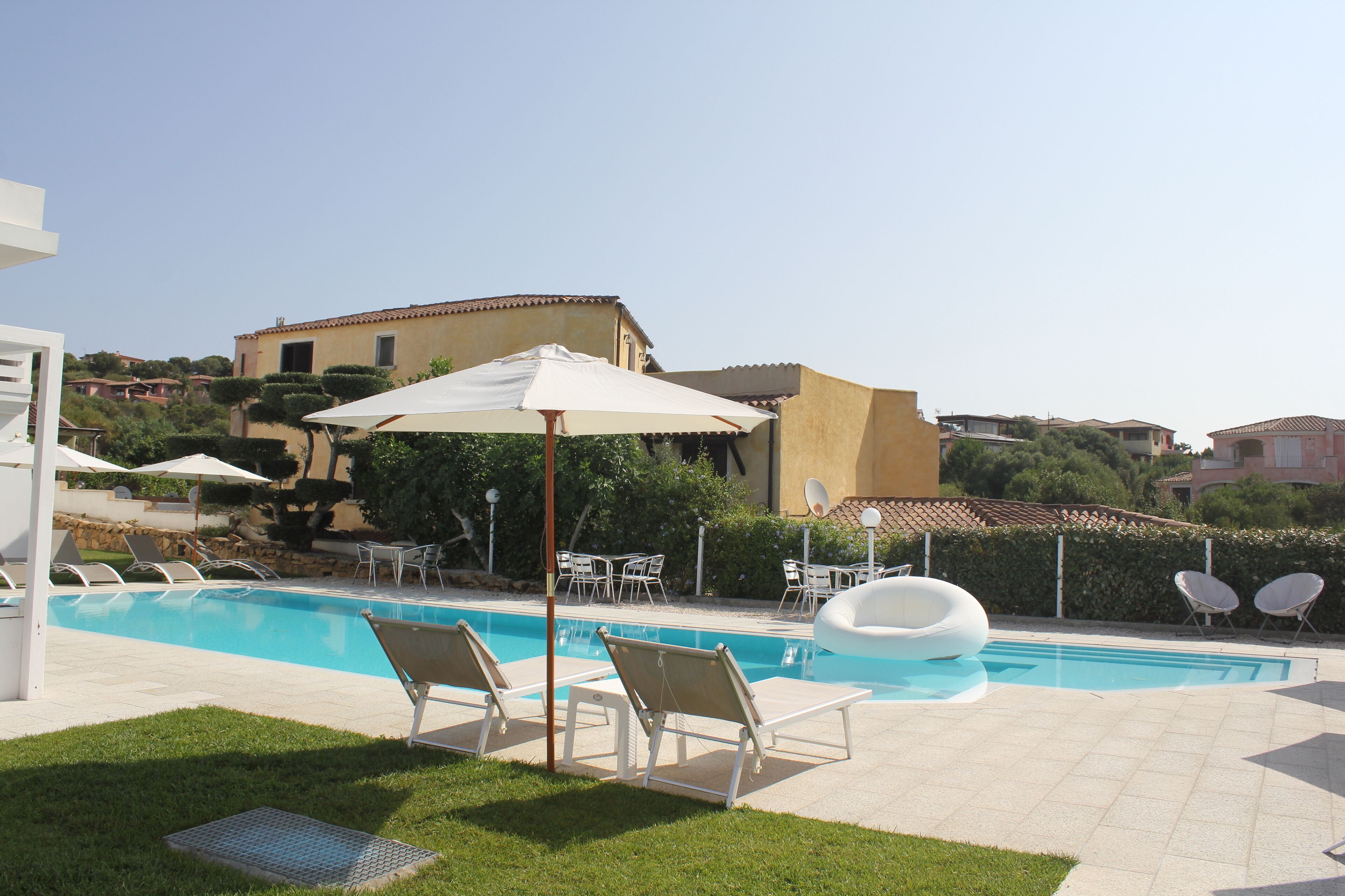 Une piscine entourée de chaises longues et de parasols, elle-même bordée d’un jardin aménagé et avec des bâtiments en arrière-plan.