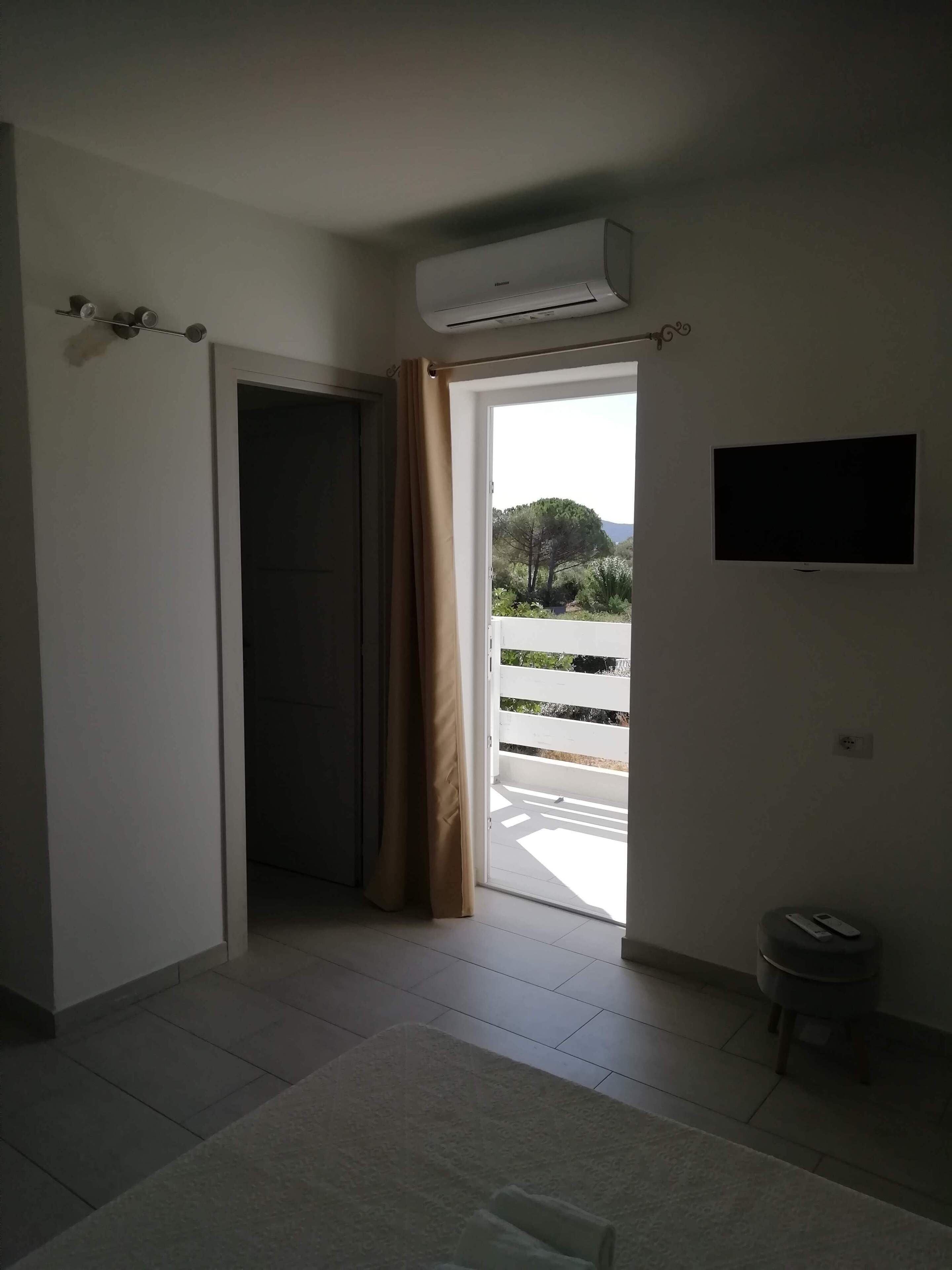 Una habitación con puerta corrediza que da a un balcón, aire acondicionado instalado, televisor de pantalla plana y un taburete pequeño.