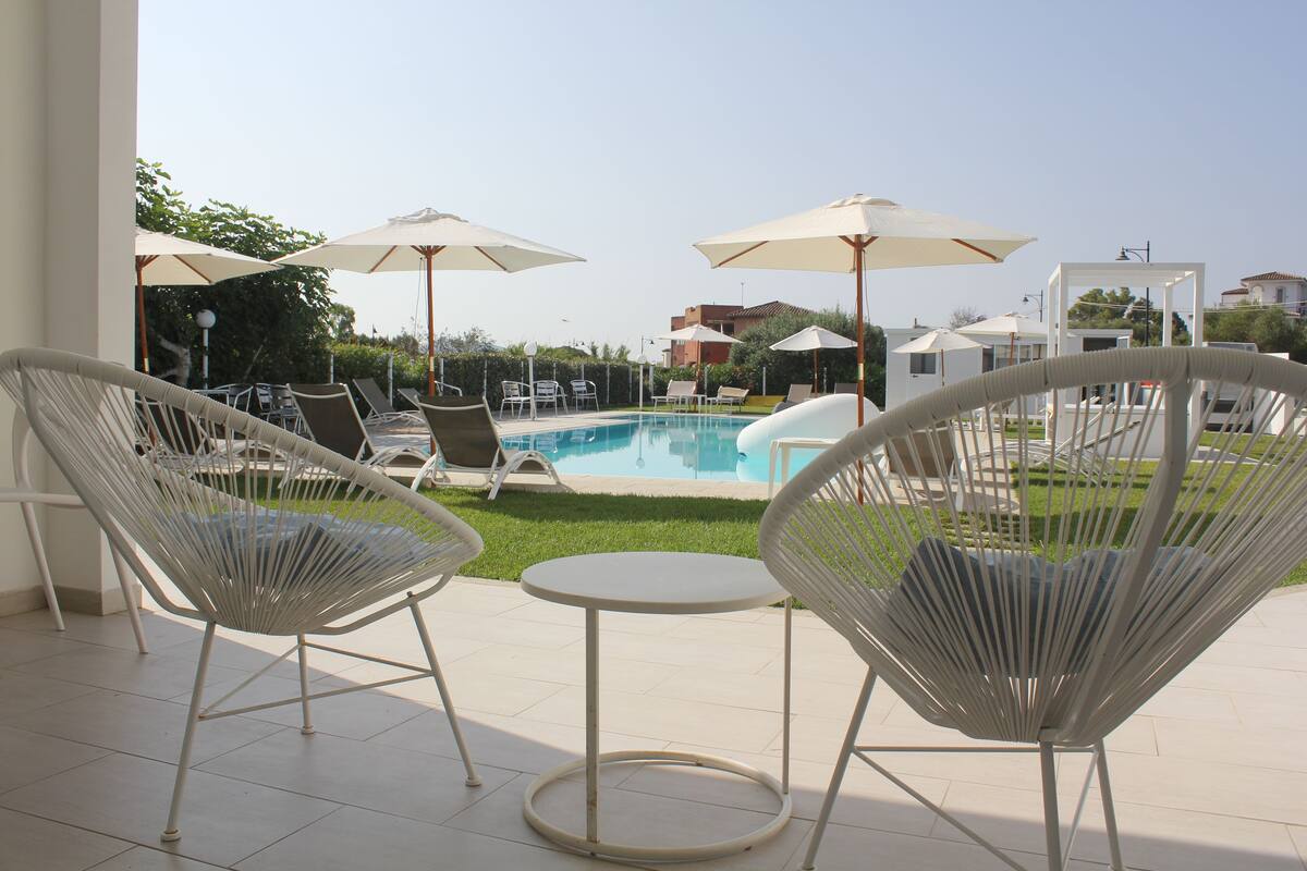 A poolside area with white wicker chairs, a small round table, and umbrellas providing shade.