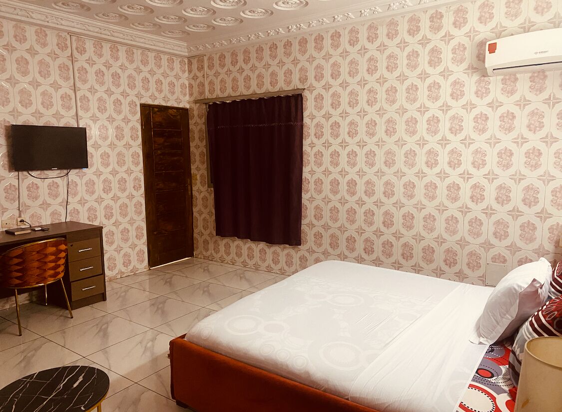 A bedroom with a bed, a desk, a chair, a TV, and a window with curtains.