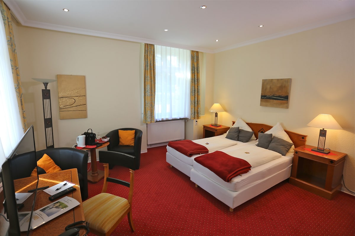 Een hotelkamer met een tweepersoonsbed, twee nachtkastjes met lampen, een bureau met stoel, een kleine tafel met stoel en een raam met gordijnen.