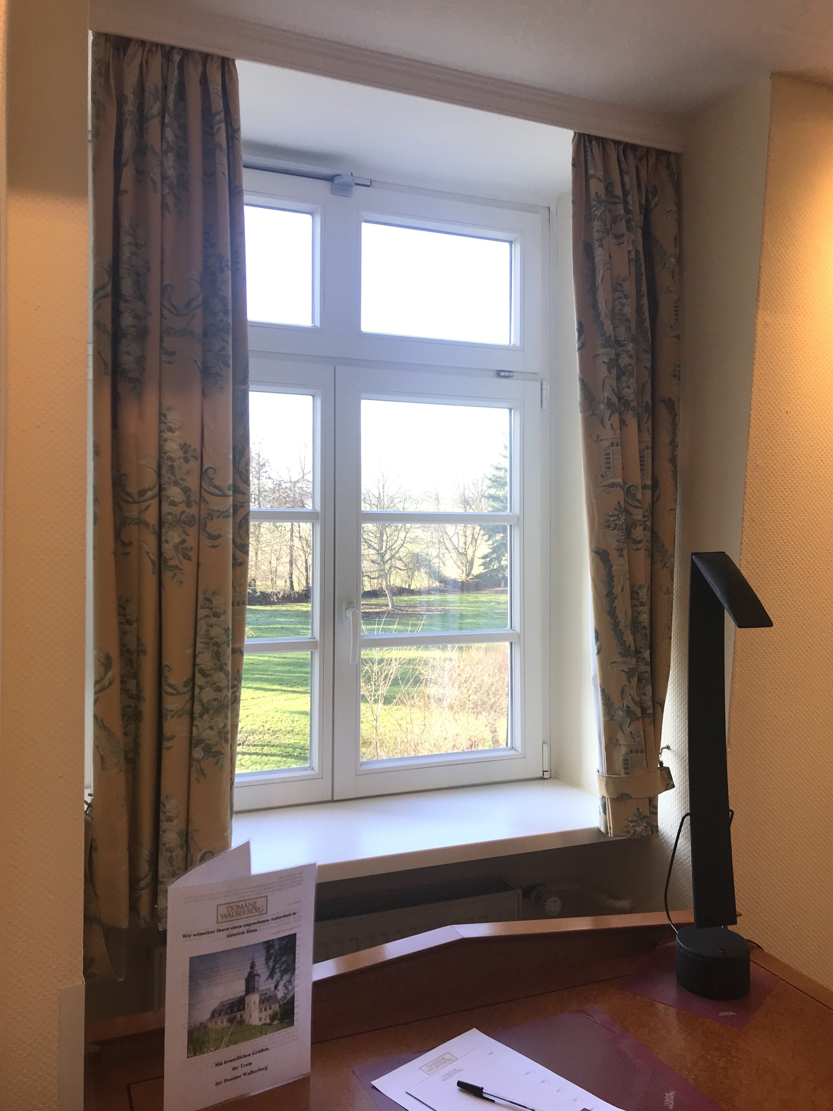 Una habitación con ventana, cortinas, un escritorio con lámpara y un folleto en el alféizar.