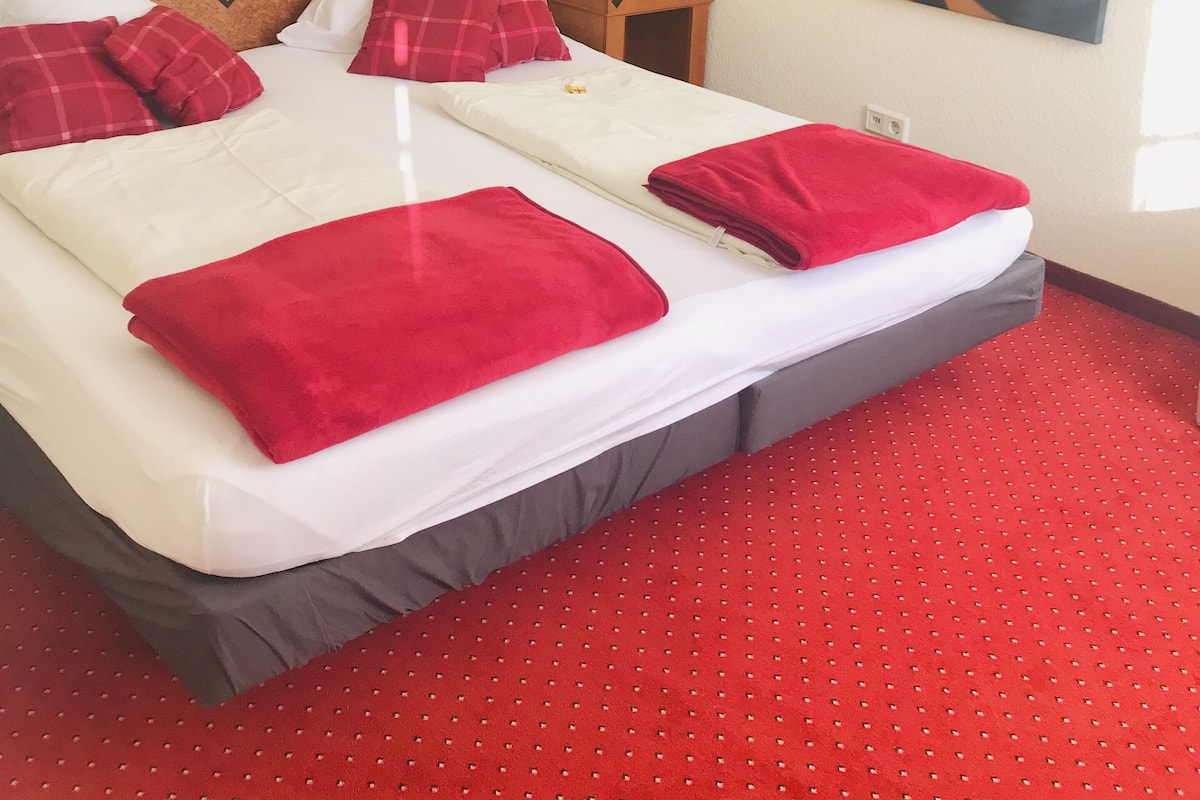 Een tweepersoonsbed met rood-witte beddengoed, een houten hoofdbord, een vloer met rode vloerbedekking en twee kunstwerken aan de muur.