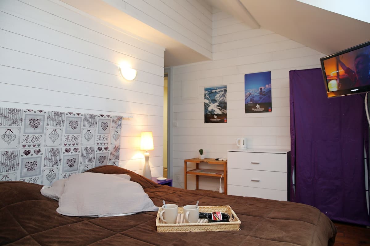A hotel room with a bed, a bedside table with a lamp, a small wooden side table, a TV, and a wall with patterned wallpaper and posters.