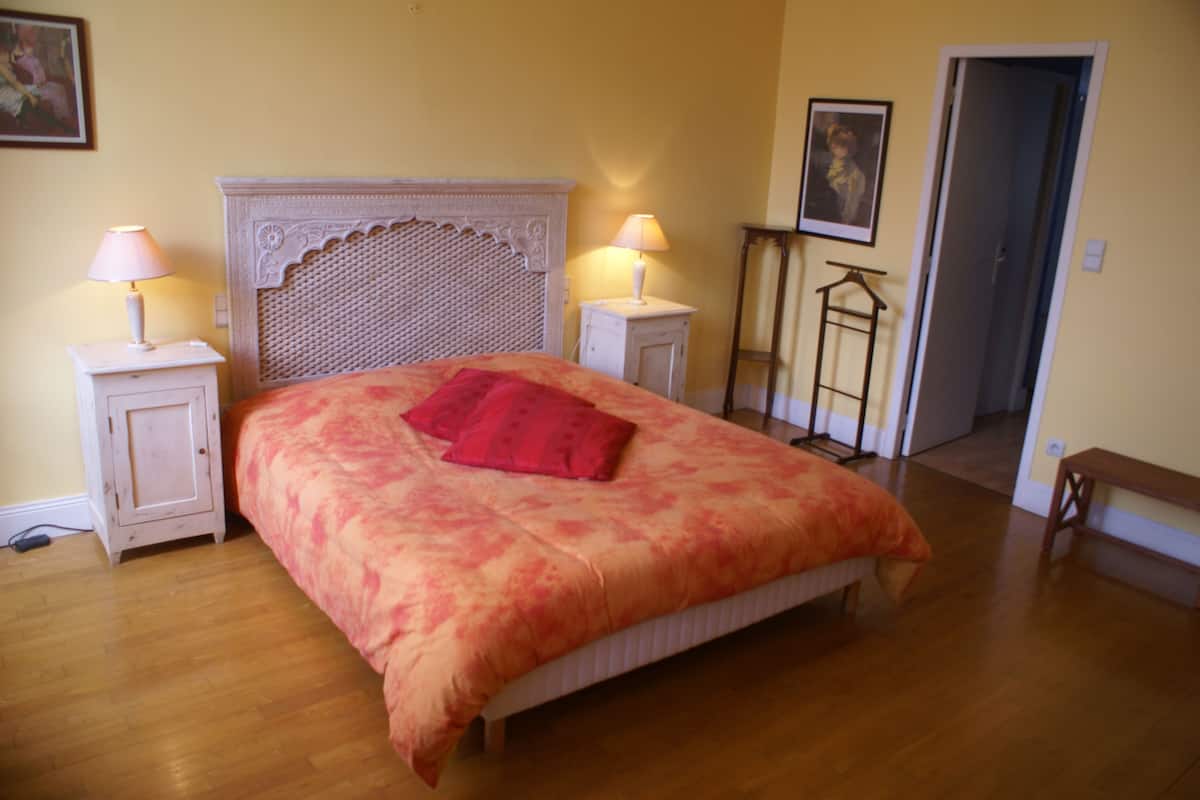 A bedroom with a large bed, two bedside tables with lamps, a wooden floor, and a door leading to another room.