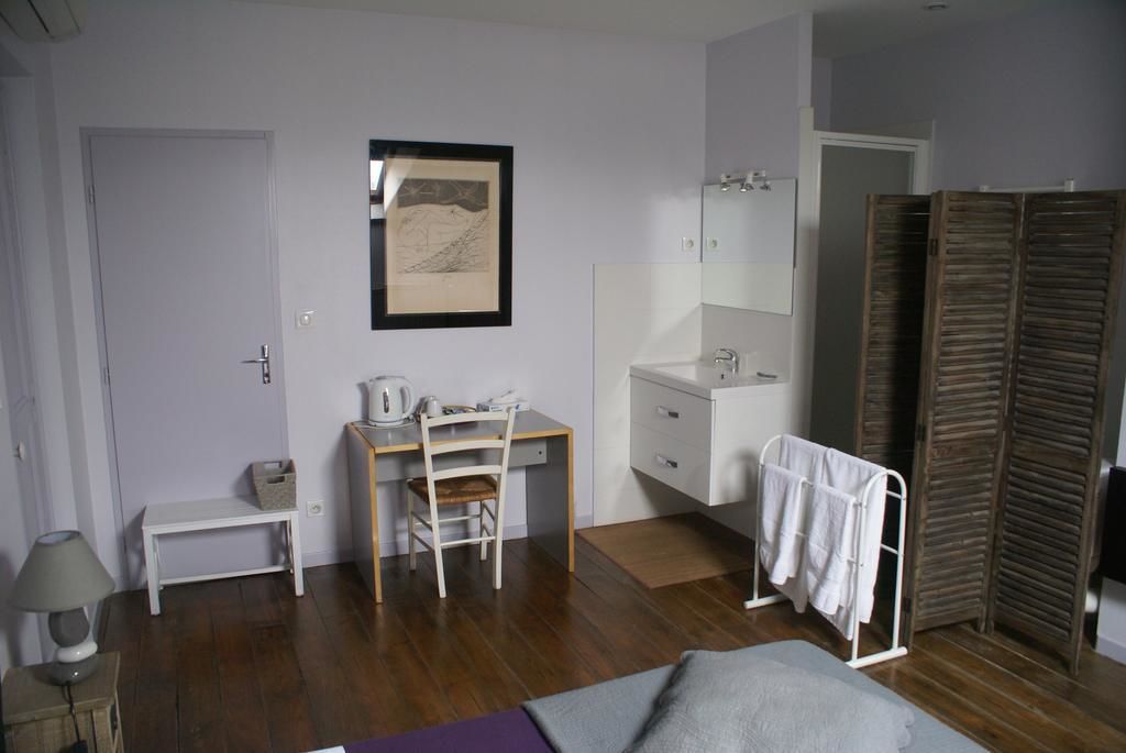 Une chambre avec un bureau en bois, une table de chevet blanche, un lit, une salle de bain avec un lavabo et un miroir, et une armoire avec des volets en bois.