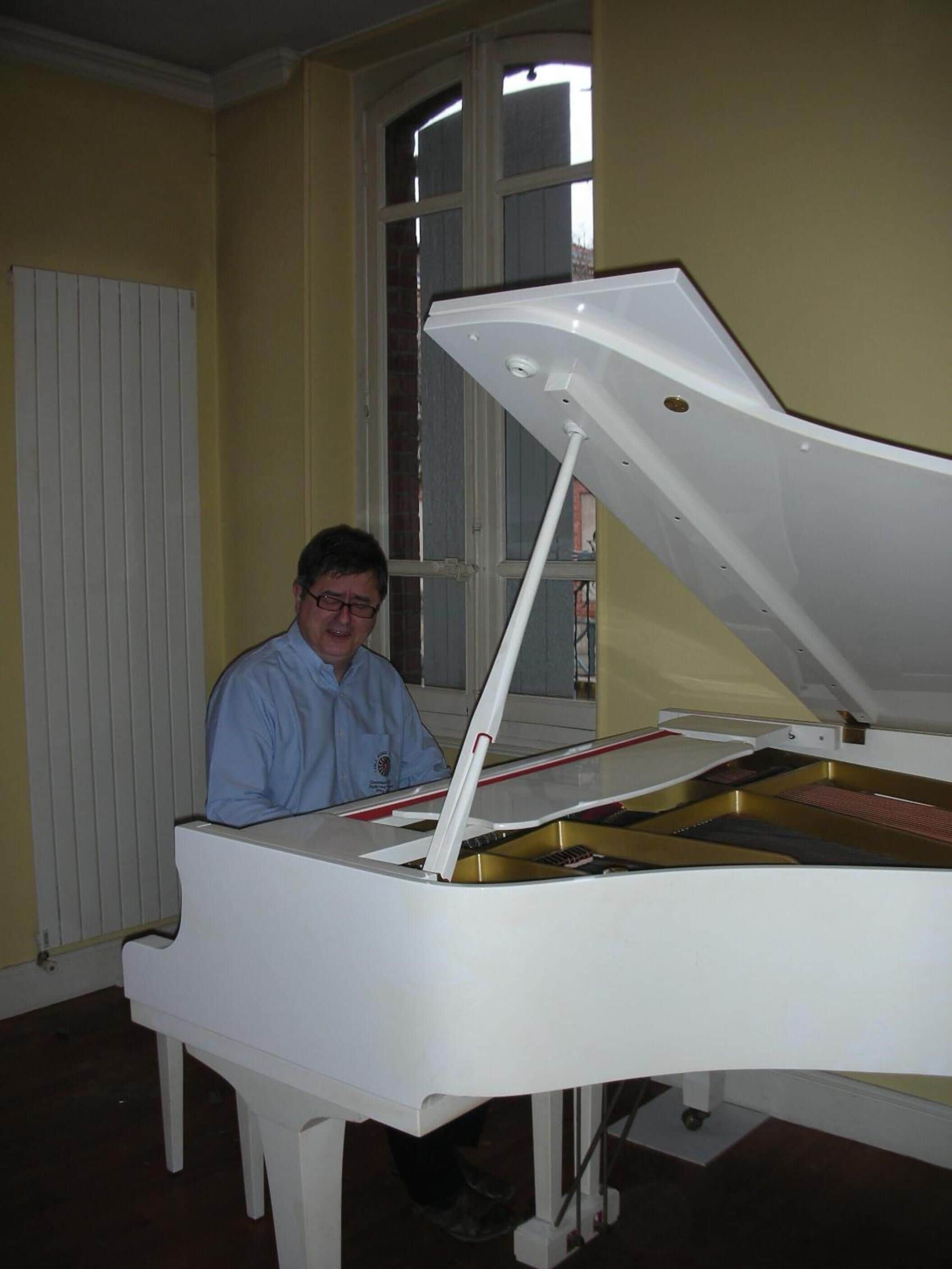 A man seated at a white grand piano in a room with yellow walls and a window.