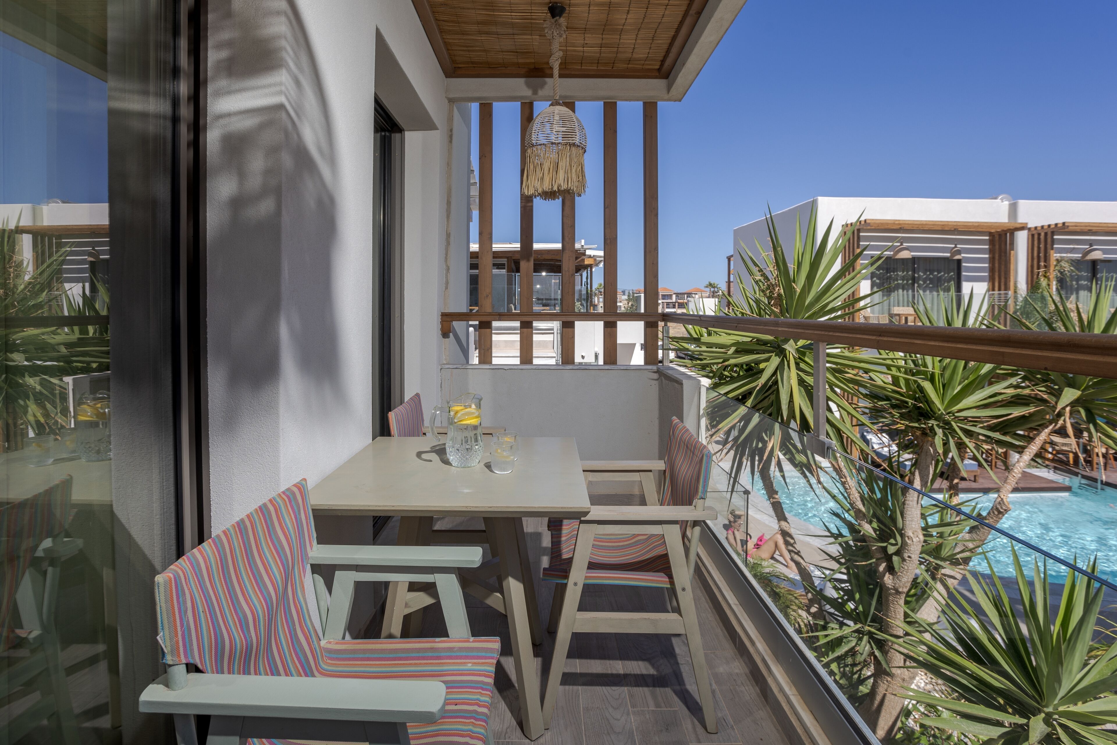 A balcony with a table and chairs, a hanging light fixture, and a view of a swimming pool.