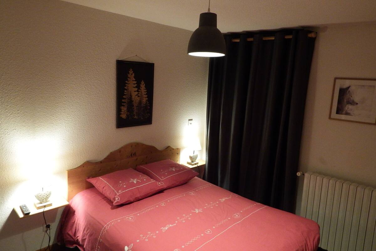 Une chambre à coucher avec une tête de lit en bois, une couvre-lit rouge, une table de chevet avec une lampe, un tableau encadré au mur et une fenêtre avec des rideaux foncés.