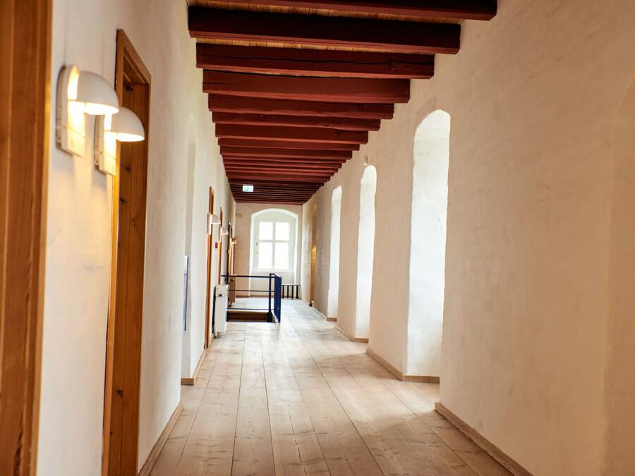 Ein Korridor mit Holzbalken, weißen Wänden und Bogenfenstern.