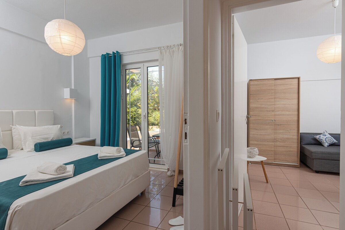 A hotel room with a bed, a small table, a sofa, and a sliding door leading to an outdoor area.