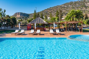 A swimming pool with lounge chairs and a shaded area, surrounded by greenery and a mountainous backdrop. - W Suites by Estia (Hersonissos)