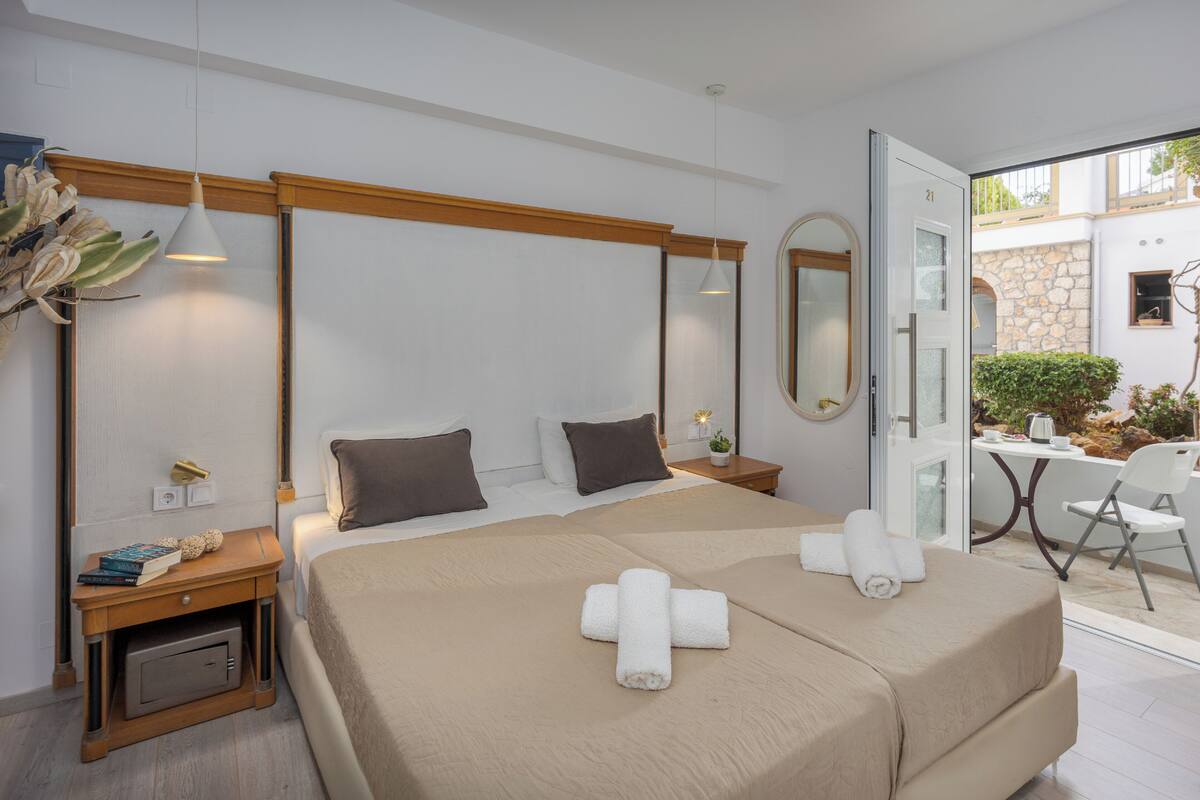 A hotel room with a bed, bedside tables, a mirror, a small table with a chair, and a view of the outside.