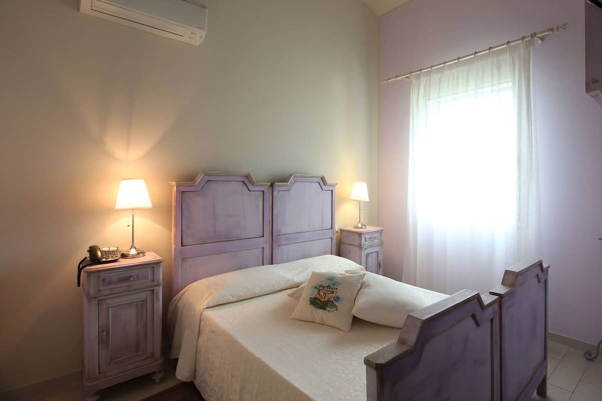 A bedroom with a bed, two bedside tables with lamps, a nightstand, a window with curtains, and a wall-mounted air conditioner.