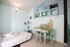 Un dormitorio con cama, un escritorio pequeño con silla, una estantería con adornos y una repisa con un radio.