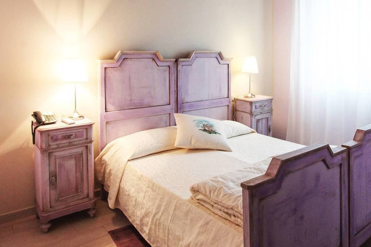 A bedroom with a bed, bedside tables, a nightstand, a lamp, a window with curtains, and an air conditioning unit.