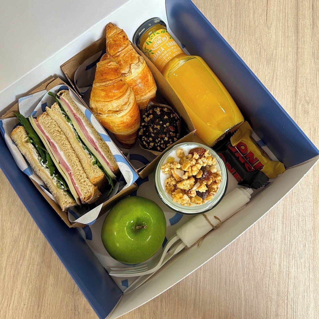 Eine Essbox mit Sandwiches, einem Croissant, einem Apfel, einem Glas Orangensaft, einem Keks und einer Müsliriegel.