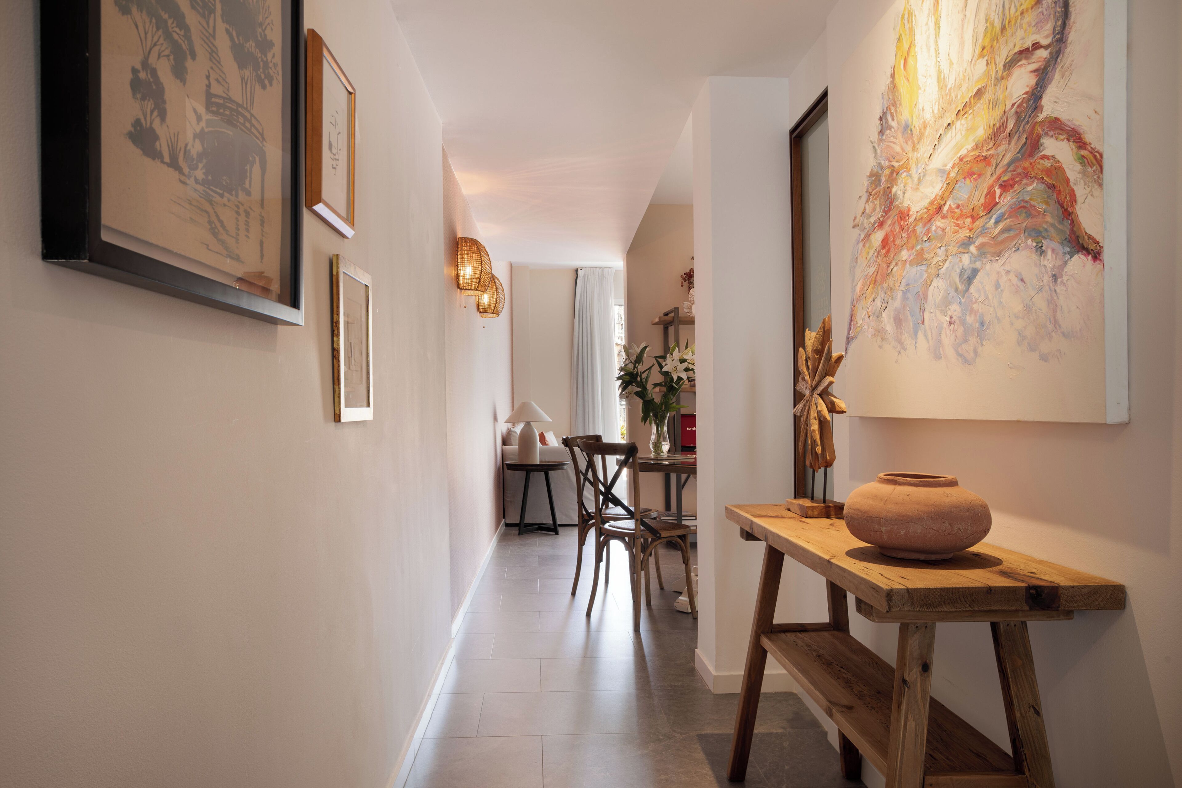 A hallway with framed artwork, a wooden table with a vase, and a dining area in the background.