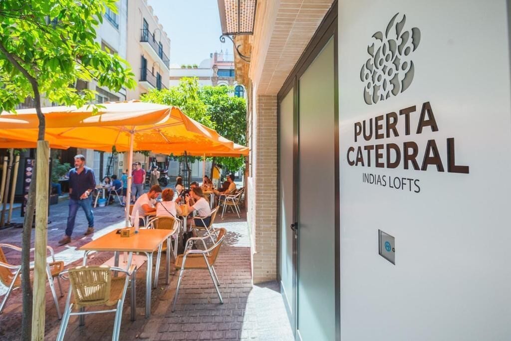 An outdoor seating area with tables and chairs, a building entrance with a sign reading 'PUERTA CATEDRAL INDIAS LOFTS,' and a tree in the background.