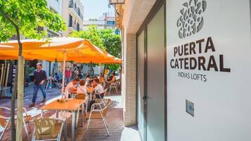 An outdoor seating area with tables and chairs, a building entrance with a sign reading 'PUERTA CATEDRAL INDIAS LOFTS,' and a tree in the background.