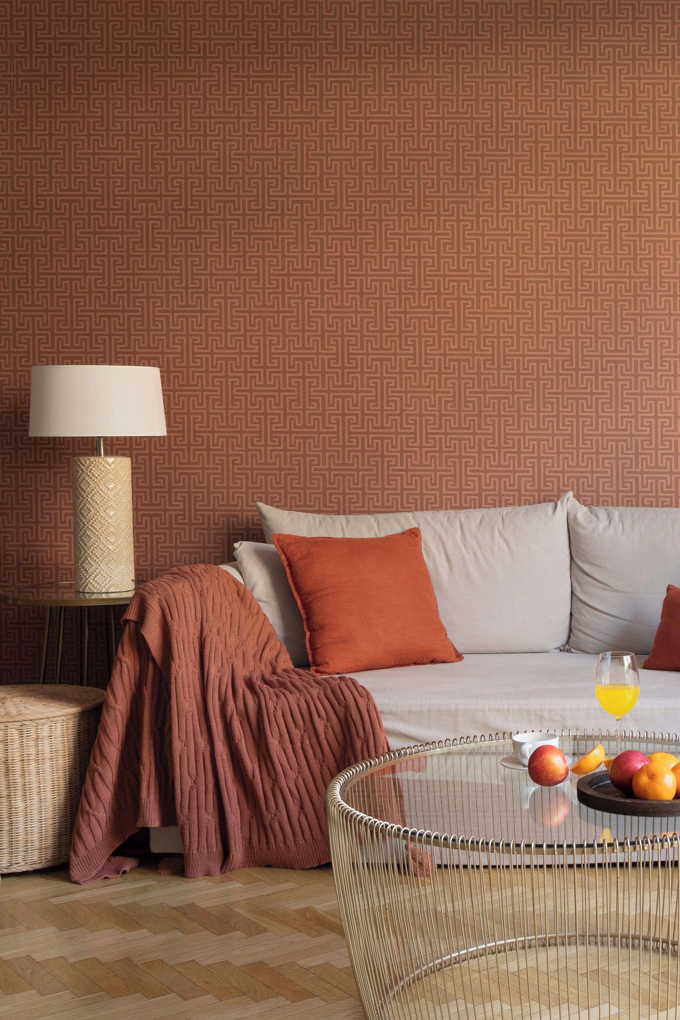 A hotel room with a patterned wallpaper, a bed with pillows, a bedside lamp, a wicker chair, a table with fruits and a glass of juice, and a woven ottoman.
