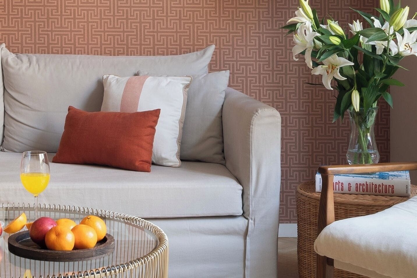 A living room with a sofa, pillows, a fruit bowl, a glass of juice, and a vase with flowers.