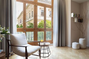 A room with a large window, a chair, a small table, a bookshelf, and a decorative branch. - Puerta Catedral Indias Lofts (Seville)