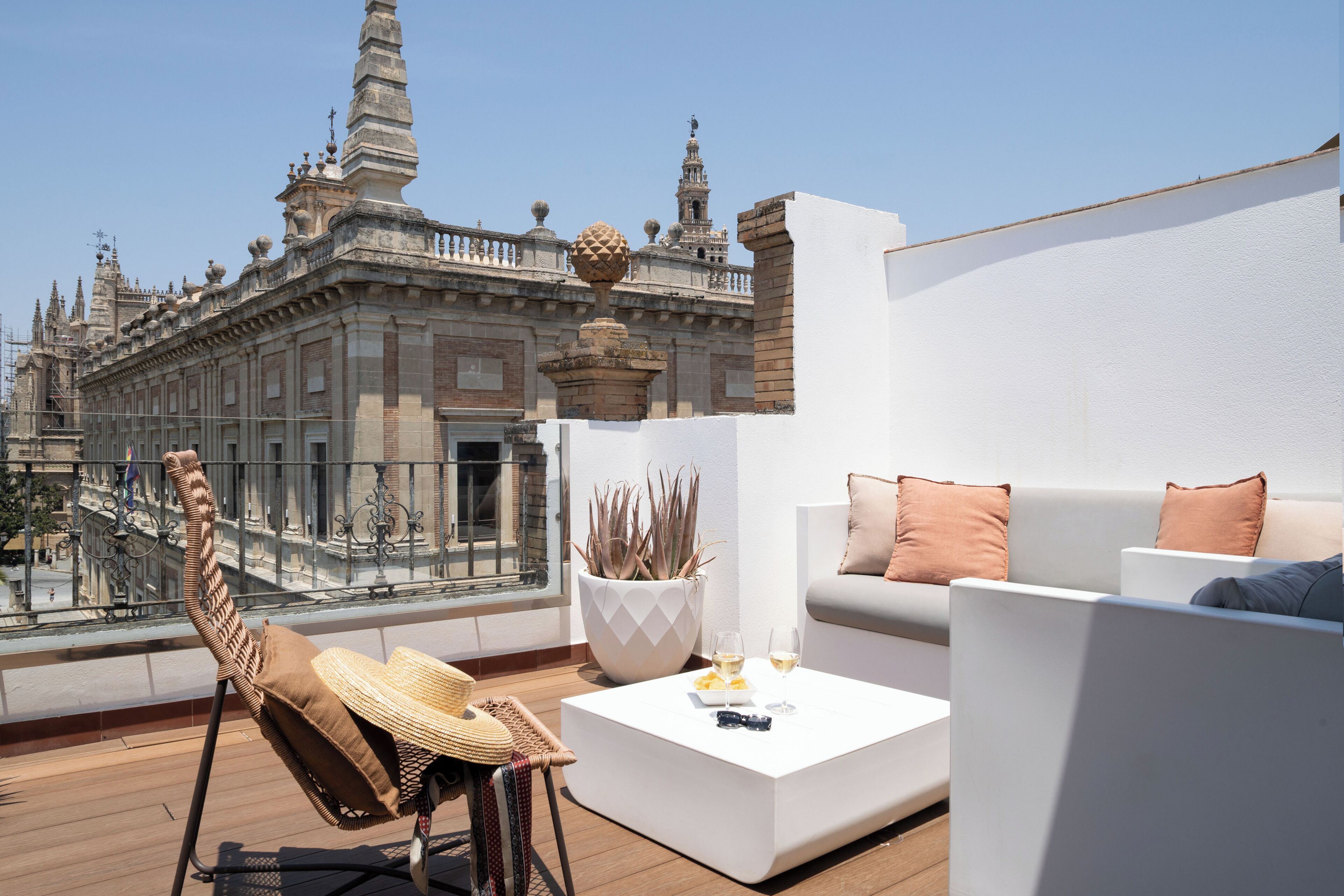 A rooftop terrace with a white sofa, a wicker chair, and a potted plant. A balustrade overlooks a historic building with ornate architecture.