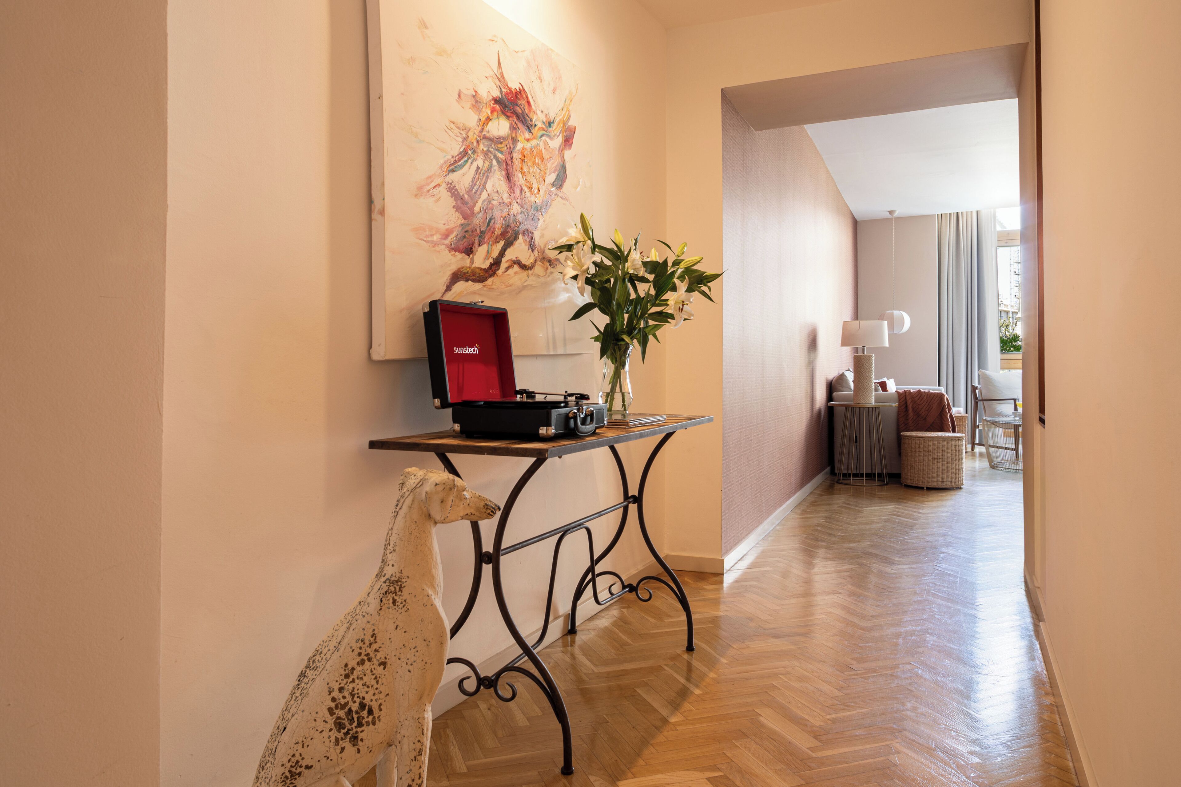 A hallway with a wooden floor, a table with a record player, a vase with flowers, and a painting on the wall.