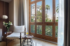 A room with a large window, a chair, a small table, and a vase with flowers. - Puerta Catedral Indias Lofts (Seville)