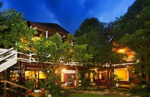 Une maison traditionnelle en bois, dotée d’un patio couvert, entourée d’une végétation luxuriante et agrémentée de plantes en pot.