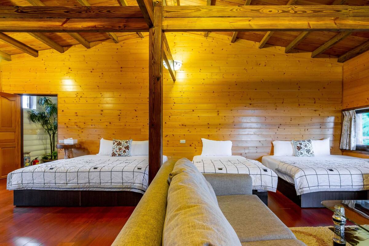 Ein blockhausähnliches Zimmer mit Holzwänden und -decke, zwei Betten mit weißer Bettwäsche, einer Couch und einem Fenster mit Vorhängen.