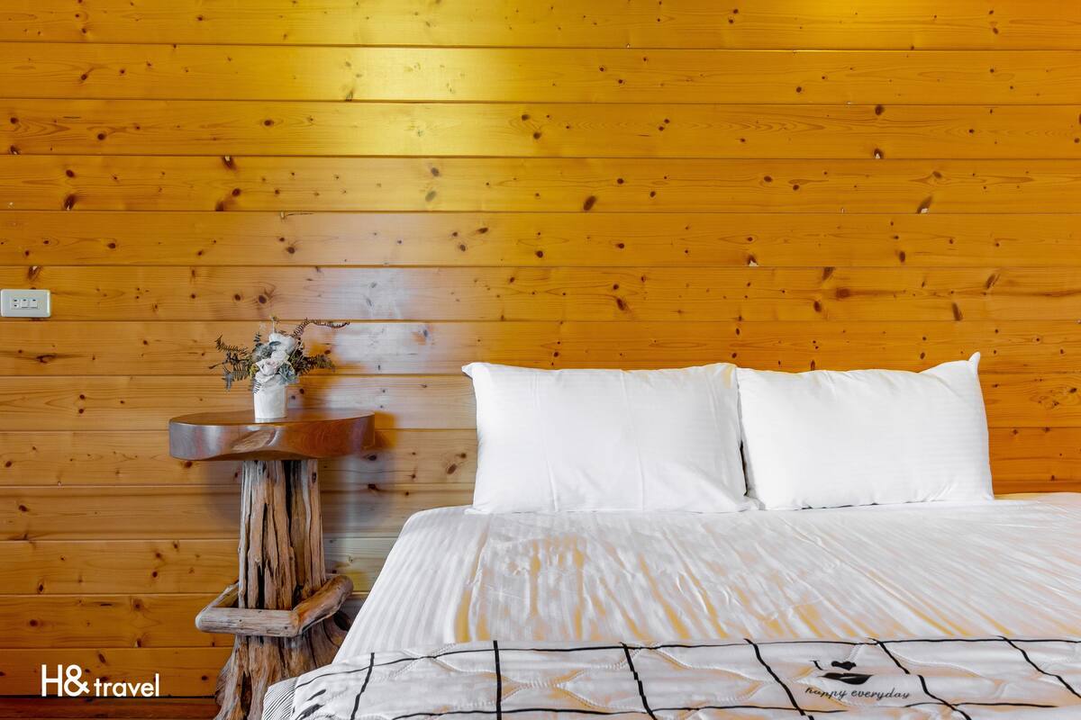 Ein Zimmer mit Holzwandverkleidung, einem Bett, weißer Bettwäsche, einem kleinen Holztisch mit einer Vase und einem Schild mit der Aufschrift "H& travel".